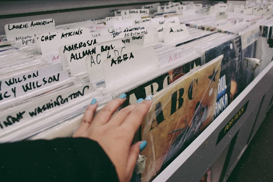 A hand with painted nails browsing through a collection of vinyl records in a store. Various artist names are written on white dividers, including 'Laurie Anderson', 'Ambrosia', 'Allman Bros.' and 'ABBA'. The records are stacked vertically in a long row, and there is a sense of nostalgia and exploration in the setting.