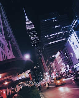 A bustling city street at night with tall, brightly lit skyscrapers. The iconic Empire State Building is prominently visible against the night sky. The street is filled with traffic, including a yellow taxi, and people walking on the sidewalks. Neon signs and illuminated billboards add vibrant colors to the scene, suggesting a lively urban environment.