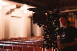 A professional film camera is set up on a tripod in an empty auditorium, capturing rows of wooden seats. A person wearing a dark shirt and a cap is standing next to the camera, possibly operating it.