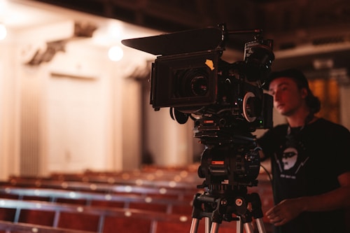 A professional film camera is set up on a tripod in an empty auditorium, capturing rows of wooden seats. A person wearing a dark shirt and a cap is standing next to the camera, possibly operating it.