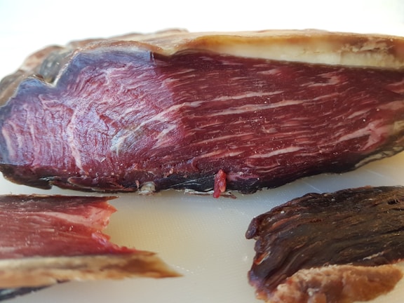 A close-up view of a piece of cured meat, displaying the marbled texture and dark reddish-brown color. The cut reveals intricate patterns of fat interwoven with the meat fibers, highlighting the artisanal preparation.