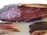 A close-up view of a piece of cured meat, displaying the marbled texture and dark reddish-brown color. The cut reveals intricate patterns of fat interwoven with the meat fibers, highlighting the artisanal preparation.
