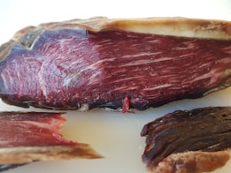 A close-up view of a piece of cured meat, displaying the marbled texture and dark reddish-brown color. The cut reveals intricate patterns of fat interwoven with the meat fibers, highlighting the artisanal preparation.