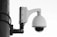 A security camera is mounted on a pole with a large, dome-shaped cover. The structure is composed of metal and plastic with visible screws and mounting hardware. Black cables are connected to the device, and the overall design suggests a robust surveillance setup.