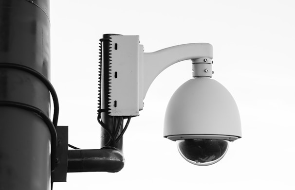 A security camera is mounted on a pole with a large, dome-shaped cover. The structure is composed of metal and plastic with visible screws and mounting hardware. Black cables are connected to the device, and the overall design suggests a robust surveillance setup.