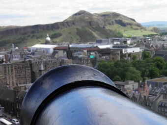 A large, weathered cannon barrel is prominently featured in the foreground, overlooking a cityscape with historic buildings. In the background, a dome structure is visible among the city structures, and a distinctive hill with rocky outcrops rises prominently, covered in green foliage and lined with paths.