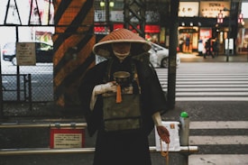 A person wearing traditional attire, including a wide-brimmed hat, stands on a street. The background features urban elements such as a zebra crossing, a busy street with cars, and storefronts. The person's attire includes a robe and white gloves, and they are holding a bowl and a prayer bead.
