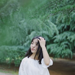 A woman with dark hair touches her hair while standing outdoors. She wears a white blouse and is surrounded by lush green trees, creating a serene and natural backdrop.