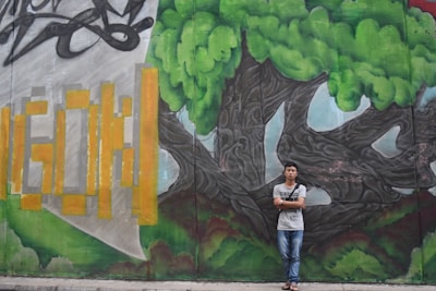 A young person stands in front of a vibrant graffiti mural featuring a large tree with lush green foliage and twisting, dark branches. To the left, the mural includes stylized yellow and orange geometric shapes against a gray and green background.