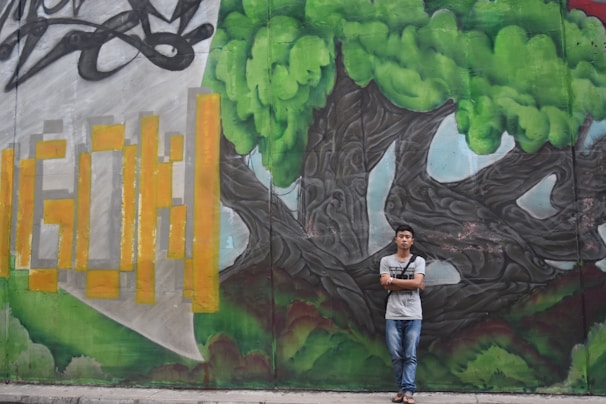 A young person stands in front of a vibrant graffiti mural featuring a large tree with lush green foliage and twisting, dark branches. To the left, the mural includes stylized yellow and orange geometric shapes against a gray and green background.