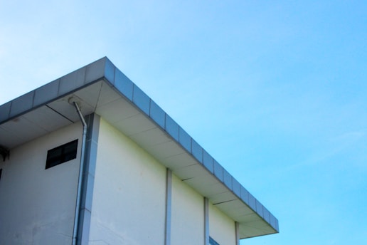 A modern building with a flat roof and a light blue sky in the background. The structure features a clean, minimalist design with a visible gutter system along its edge.