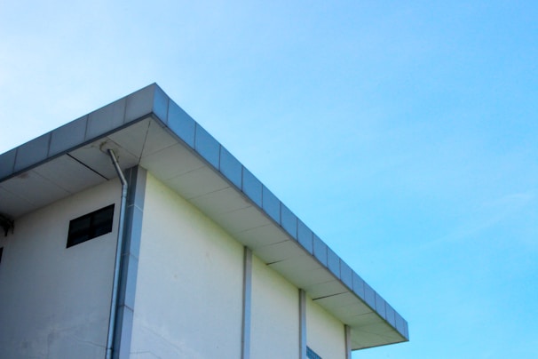 A modern building with a flat roof and a light blue sky in the background. The structure features a clean, minimalist design with a visible gutter system along its edge.