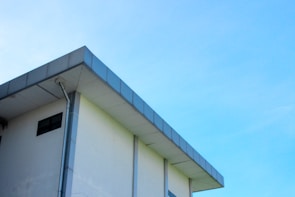 A modern building with a flat roof and a light blue sky in the background. The structure features a clean, minimalist design with a visible gutter system along its edge.