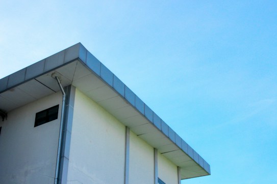 A modern building with a flat roof and a light blue sky in the background. The structure features a clean, minimalist design with a visible gutter system along its edge.