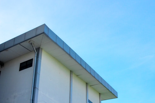 A modern building with a flat roof and a light blue sky in the background. The structure features a clean, minimalist design with a visible gutter system along its edge.