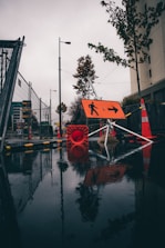 A wet urban street scene features construction barriers and signage with orange cones and a reflective wet surface. Trees line the side of the road, and a prominent orange directional sign points to the right.