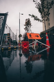 A wet urban street scene features construction barriers and signage with orange cones and a reflective wet surface. Trees line the side of the road, and a prominent orange directional sign points to the right.