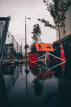 A wet urban street scene features construction barriers and signage with orange cones and a reflective wet surface. Trees line the side of the road, and a prominent orange directional sign points to the right.