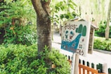 A wooden sign with colorful illustrations of children, trees, and a river, surrounded by lush green foliage. The sign features text in a foreign language and has a playful, cartoonish style.