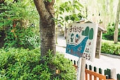 A wooden sign with colorful illustrations of children, trees, and a river, surrounded by lush green foliage. The sign features text in a foreign language and has a playful, cartoonish style.