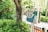 A wooden sign with colorful illustrations of children, trees, and a river, surrounded by lush green foliage. The sign features text in a foreign language and has a playful, cartoonish style.