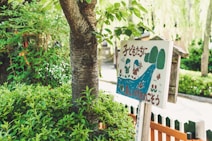 A wooden sign with colorful illustrations of children, trees, and a river, surrounded by lush green foliage. The sign features text in a foreign language and has a playful, cartoonish style.