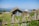 A rustic stone and wood hut with a tin roof sits in a grassy rural setting. A wooden fence surrounds the area, and other small structures with tin roofs are visible in the background. The landscape is mountainous with lush greenery and a clear blue sky.