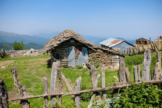 A rustic stone and wood hut with a tin roof sits in a grassy rural setting. A wooden fence surrounds the area, and other small structures with tin roofs are visible in the background. The landscape is mountainous with lush greenery and a clear blue sky.