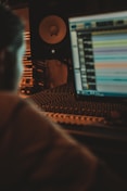 A dimly lit music production setup featuring a computer screen displaying audio editing software. In the foreground, there's a blurred silhouette of a person working at the desk. Behind the monitor, audio equipment including a large speaker and soundproofing foam are visible.