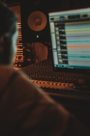 A dimly lit music production setup featuring a computer screen displaying audio editing software. In the foreground, there's a blurred silhouette of a person working at the desk. Behind the monitor, audio equipment including a large speaker and soundproofing foam are visible.