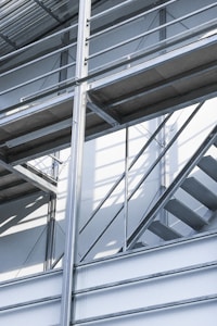 A modern architectural structure with metal beams and a stairway. The design features intersecting lines and angles, highlighted by natural lighting that creates shadows and adds depth.