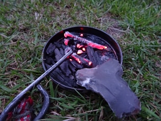 A small circular metal container holds burning charcoal pieces, with visible red embers. A stone and a metal tool are adjacent on a patch of grass.