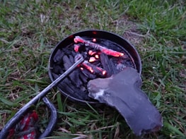 A small circular metal container holds burning charcoal pieces, with visible red embers. A stone and a metal tool are adjacent on a patch of grass.
