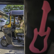 A man in a khaki uniform sits inside a black and yellow auto-rickshaw with an open side door. The vehicle is on a sunny city street, surrounded by other vehicles. In the foreground, part of the interior of another vehicle is visible, with a large pink guitar decal on a black background.