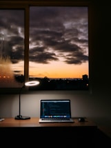 A cozy workspace featuring a wooden desk with a laptop displaying code on the screen. A modern lamp casts a circular light beside the laptop, while a serene view of a sunset with dark clouds is visible through the window behind the desk.