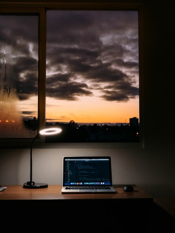 A cozy workspace featuring a wooden desk with a laptop displaying code on the screen. A modern lamp casts a circular light beside the laptop, while a serene view of a sunset with dark clouds is visible through the window behind the desk.