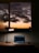 A cozy workspace featuring a wooden desk with a laptop displaying code on the screen. A modern lamp casts a circular light beside the laptop, while a serene view of a sunset with dark clouds is visible through the window behind the desk.