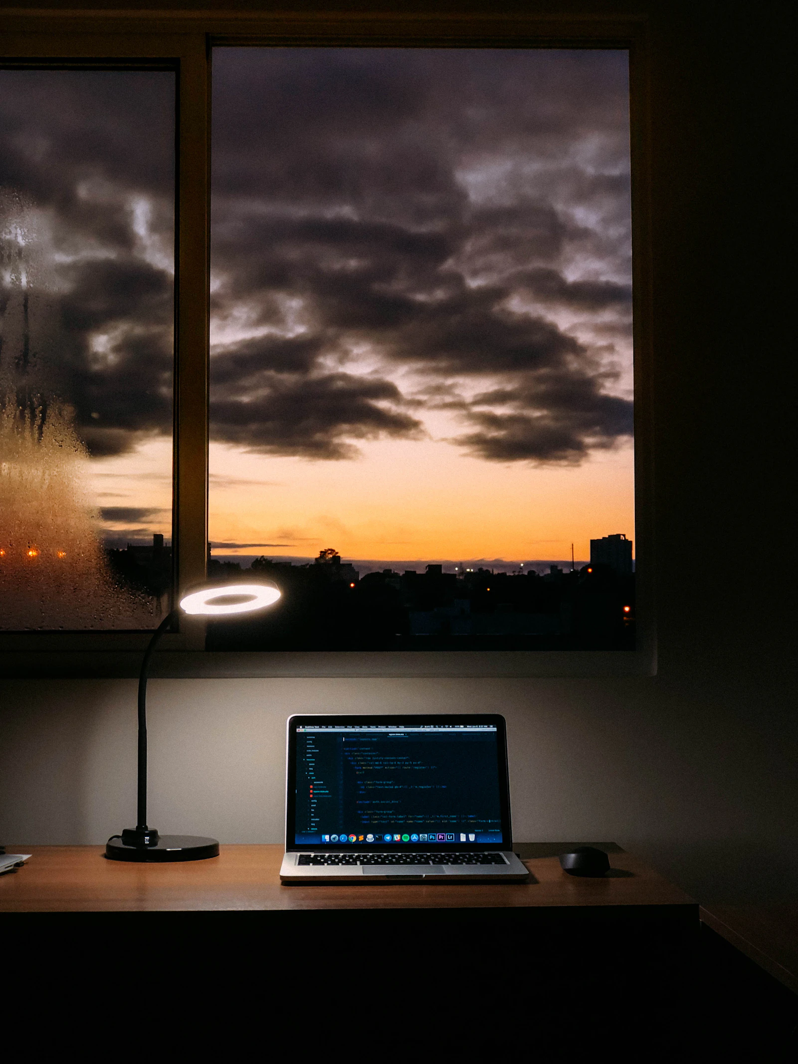 Laptop showing code on a dark desk beside a window, the screen's glow set against a moody overcast sky at first light