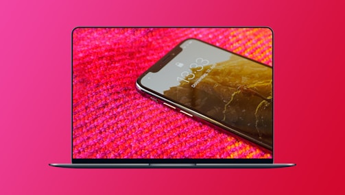 A sleek laptop screen displays a vibrant image of a smartphone positioned at an angle. The smartphone shows a lock screen with a mountain scenery wallpaper and a time display of 13:33. It rests on a textured, red and pink woven fabric, creating a contrast with its metallic frame.