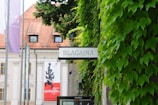 A sign with the word 'BLAGAJNA' is prominently displayed amidst lush green ivy leaves on a wall. In the background, a building with a red tiled roof and banners is visible, suggesting a historical or cultural setting.