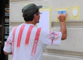 A man is painting on a wall, wearing a white shirt with red stripes and a hat. He is using a small brush on a piece of taped paper, creating an artwork that appears to depict a tree. The wall is beige and features architectural molding.