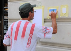 A man is painting on a wall, wearing a white shirt with red stripes and a hat. He is using a small brush on a piece of taped paper, creating an artwork that appears to depict a tree. The wall is beige and features architectural molding.
