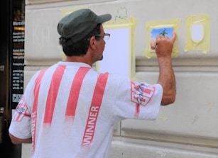A man is painting on a wall, wearing a white shirt with red stripes and a hat. He is using a small brush on a piece of taped paper, creating an artwork that appears to depict a tree. The wall is beige and features architectural molding.