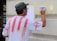 A man is painting on a wall, wearing a white shirt with red stripes and a hat. He is using a small brush on a piece of taped paper, creating an artwork that appears to depict a tree. The wall is beige and features architectural molding.