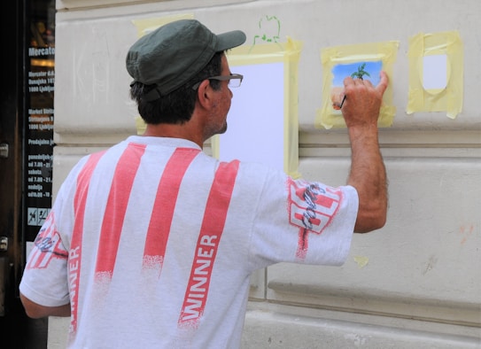 A man is painting on a wall, wearing a white shirt with red stripes and a hat. He is using a small brush on a piece of taped paper, creating an artwork that appears to depict a tree. The wall is beige and features architectural molding.