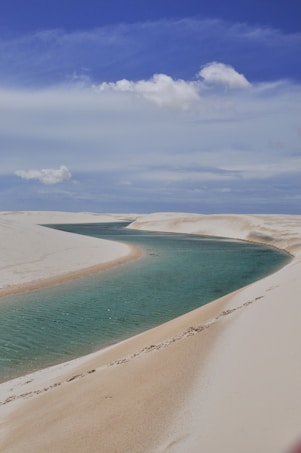 A curving turquoise lagoon is surrounded by vast white sand dunes under a mostly clear blue sky with a few scattered clouds.
