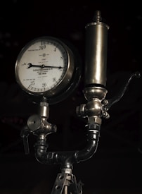 A vintage pressure gauge with a metal body and a dial marked in centimeters and kilograms per square centimeter. It features intricate metalwork and tubing, typical of an industrial device. The gauge is connected to a cylindrical component above it, possibly part of a larger machinery system.