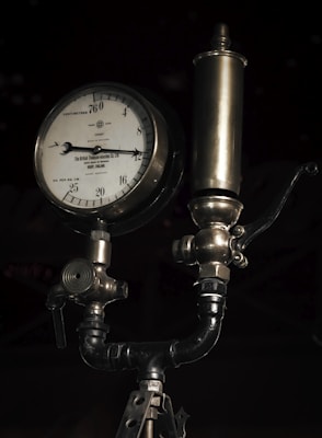 A vintage pressure gauge with a metal body and a dial marked in centimeters and kilograms per square centimeter. It features intricate metalwork and tubing, typical of an industrial device. The gauge is connected to a cylindrical component above it, possibly part of a larger machinery system.