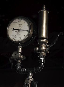 A vintage pressure gauge with a metal body and a dial marked in centimeters and kilograms per square centimeter. It features intricate metalwork and tubing, typical of an industrial device. The gauge is connected to a cylindrical component above it, possibly part of a larger machinery system.