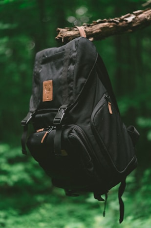 A black backpack hangs from a tree branch in a lush, green forest setting. The backpack features a leather patch and multiple compartments, with a focus on its sturdy design.